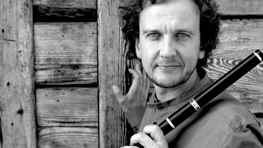Mężczyzna w jednej dłoni trzyma instrument dęty, druga dłoń spoczywa na drewnianej ścianę. Mężczyzna uśmiecha się, patrzy wprost w obiektyw.